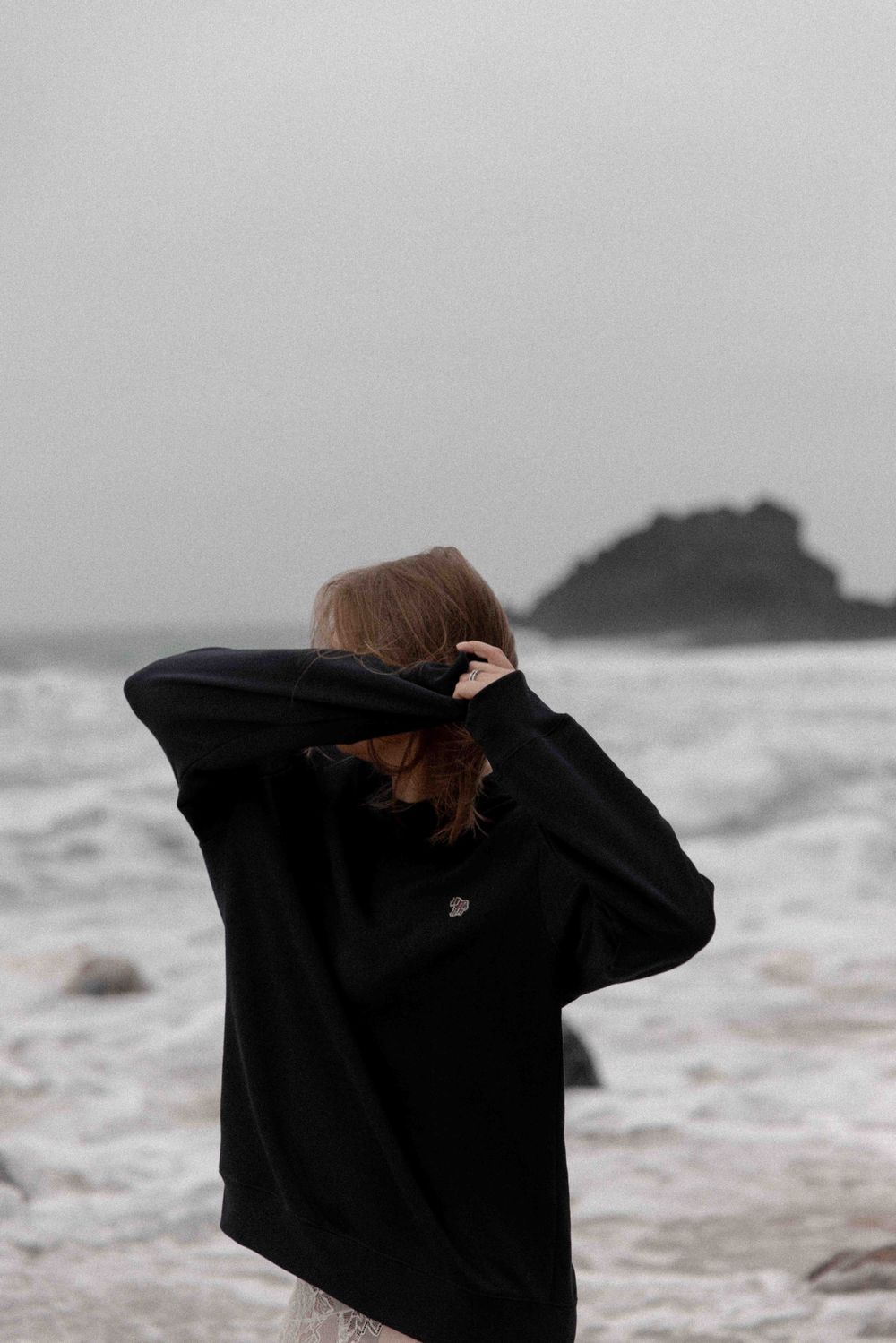 Person in black sweater shields from wind on rocky beach with waves crashing behind.