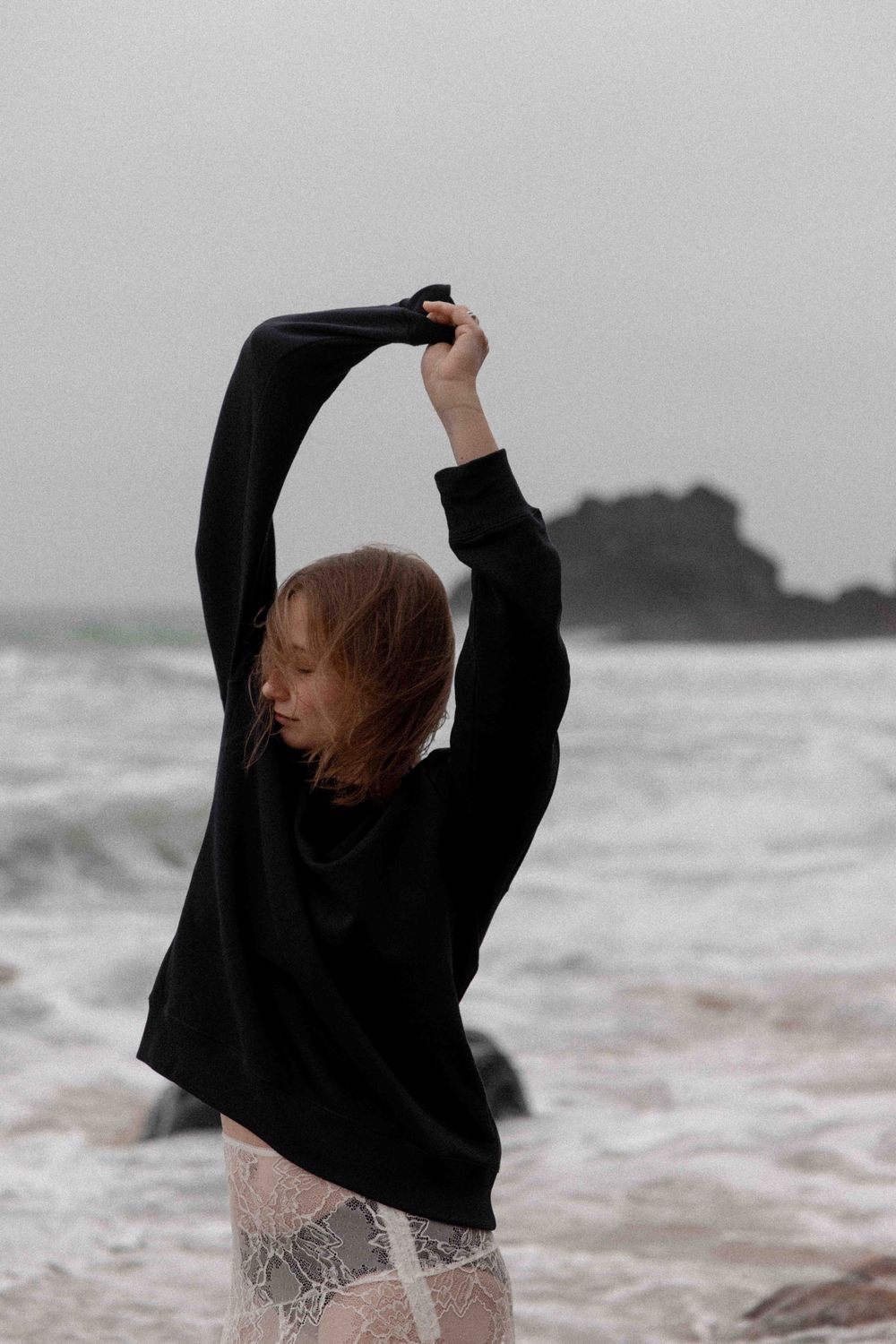 Person in black sweater stretches arms upward against moody coastal backdrop.