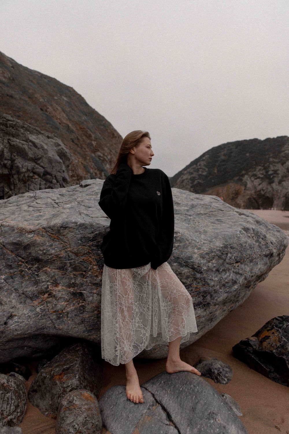 Figure in black sweater and white skirt stands barefoot on coastal rocks during overcast day.