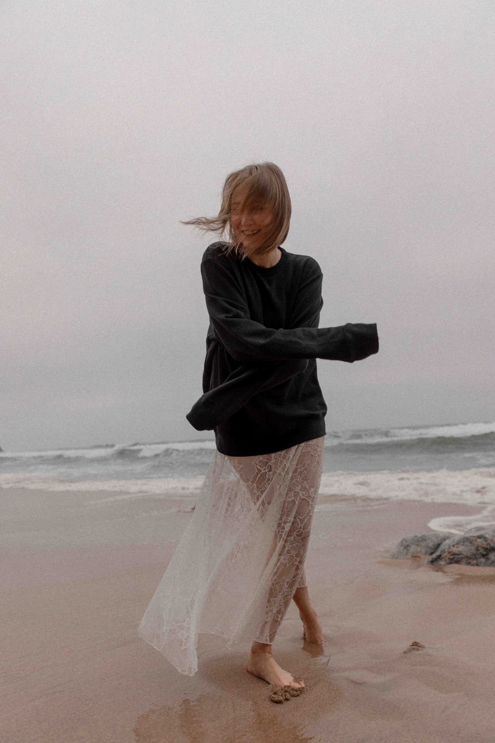 Person in black sweater and lace skirt dances on misty beach with waves in background.