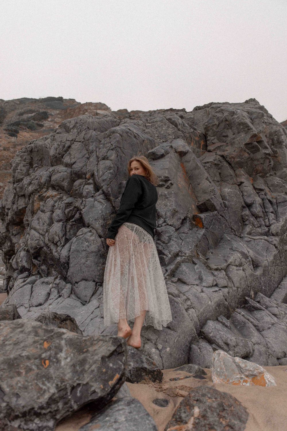 Figure in dark sweater and light skirt poses against dramatic coastal rock formation.