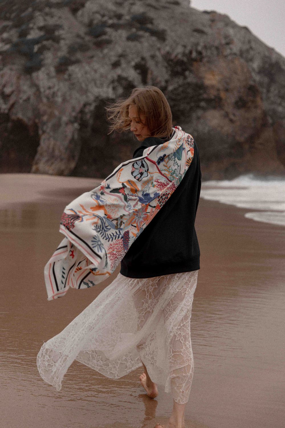 Person in floral kimono and white lace dress twirls on misty beach with rocky cliffs in background.
