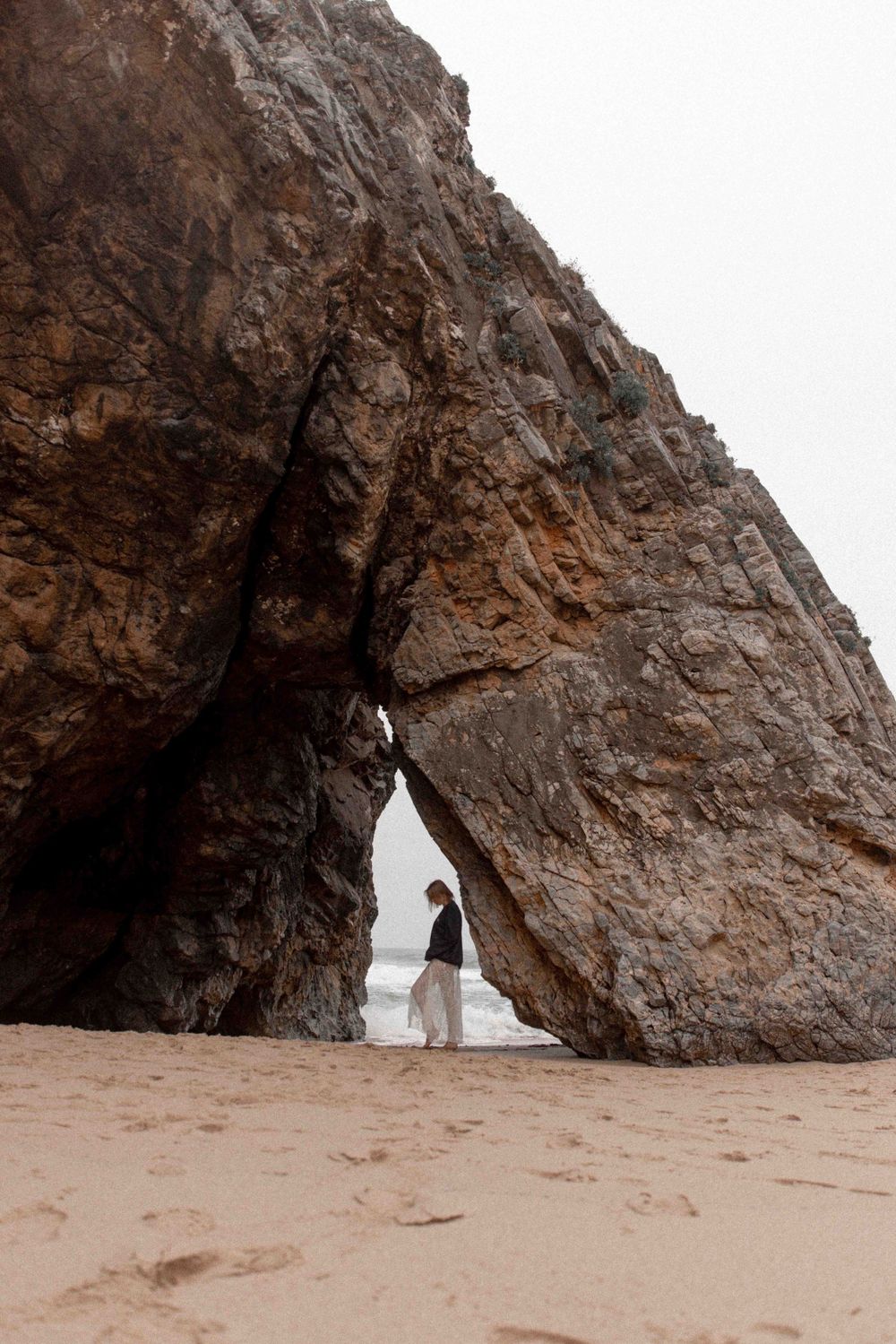 A figure stands beneath a dramatic natural stone arch formation on a sandy beach at twilight.