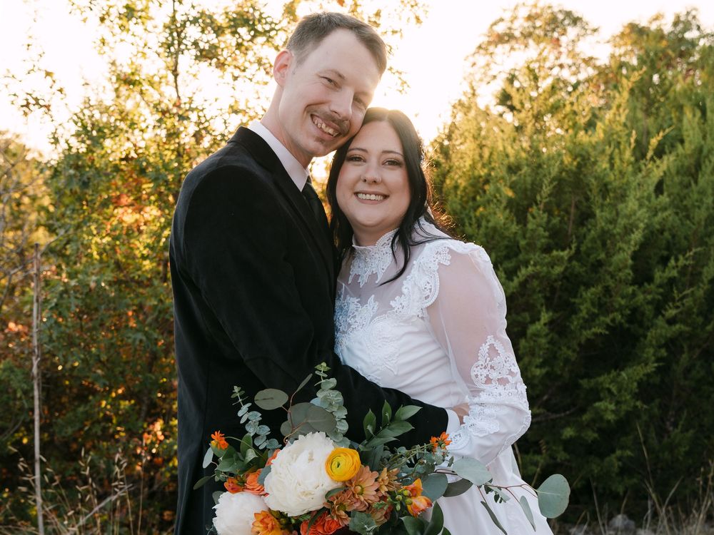 Smiling bride and groom pose outdoors at golden hour, bride holding colorful bouquet with white, orange, and yellow flowers.