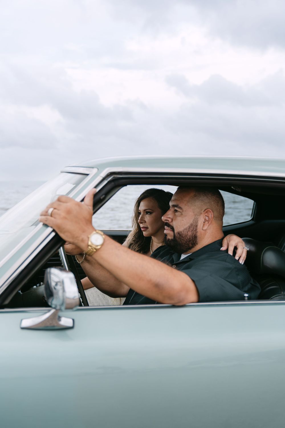 Couple sitting in a vintage mint green car near the ocean, man wearing a gold watch with arm around woman.
