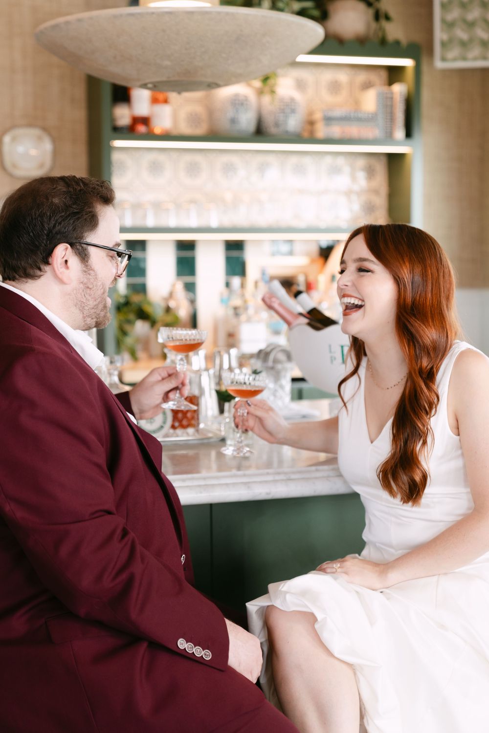 A laughing bride in white dress clinks cocktail glasses with groom in burgundy suit at a stylish bar.