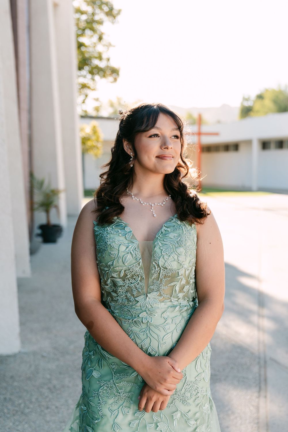 Young woman in elegant mint green lace gown with crystal necklace poses outdoors in golden hour light.