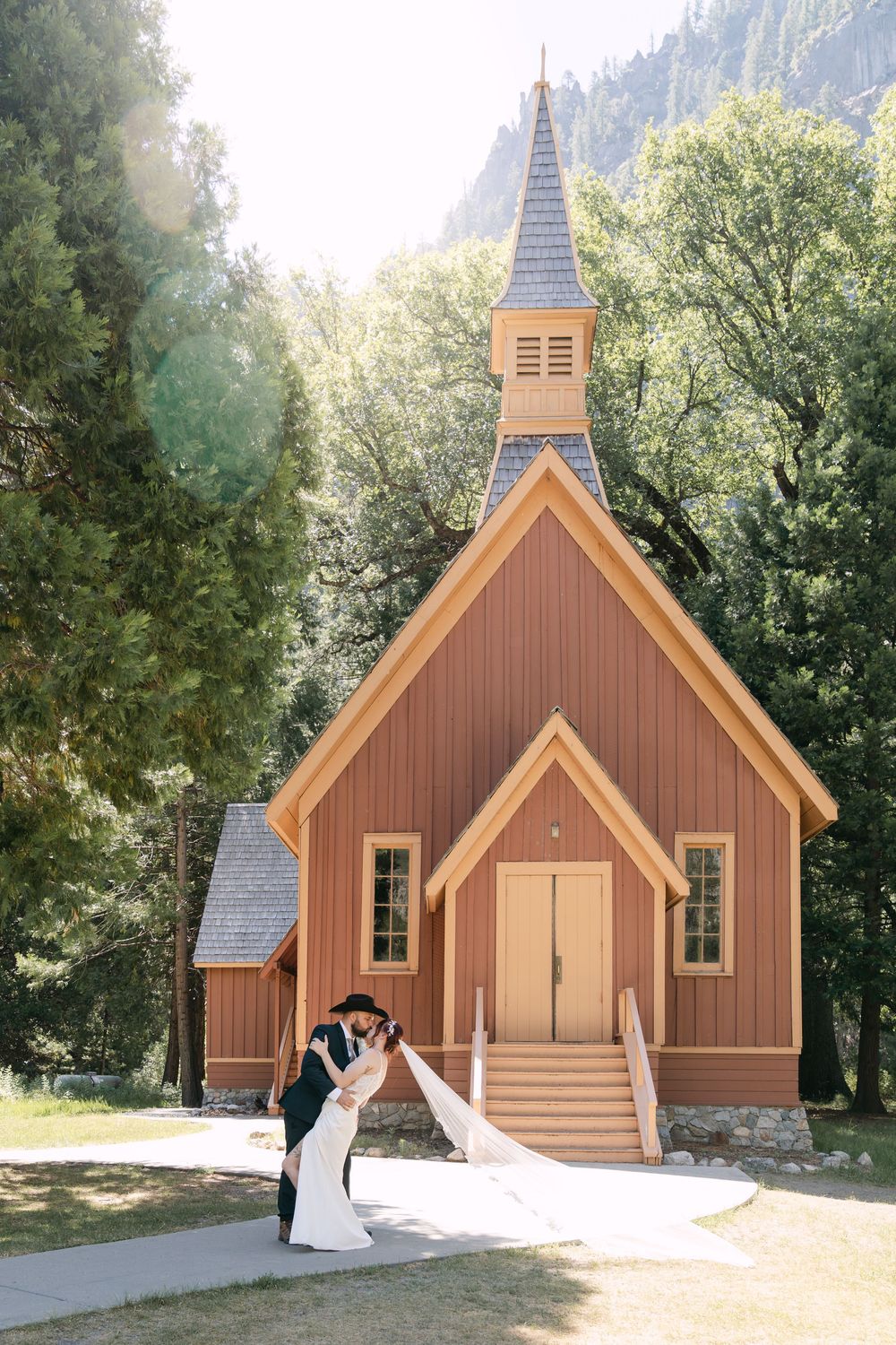 Bride and groom share a romantic dip kiss in front of a charming wooden chapel in Yosemite National Park.