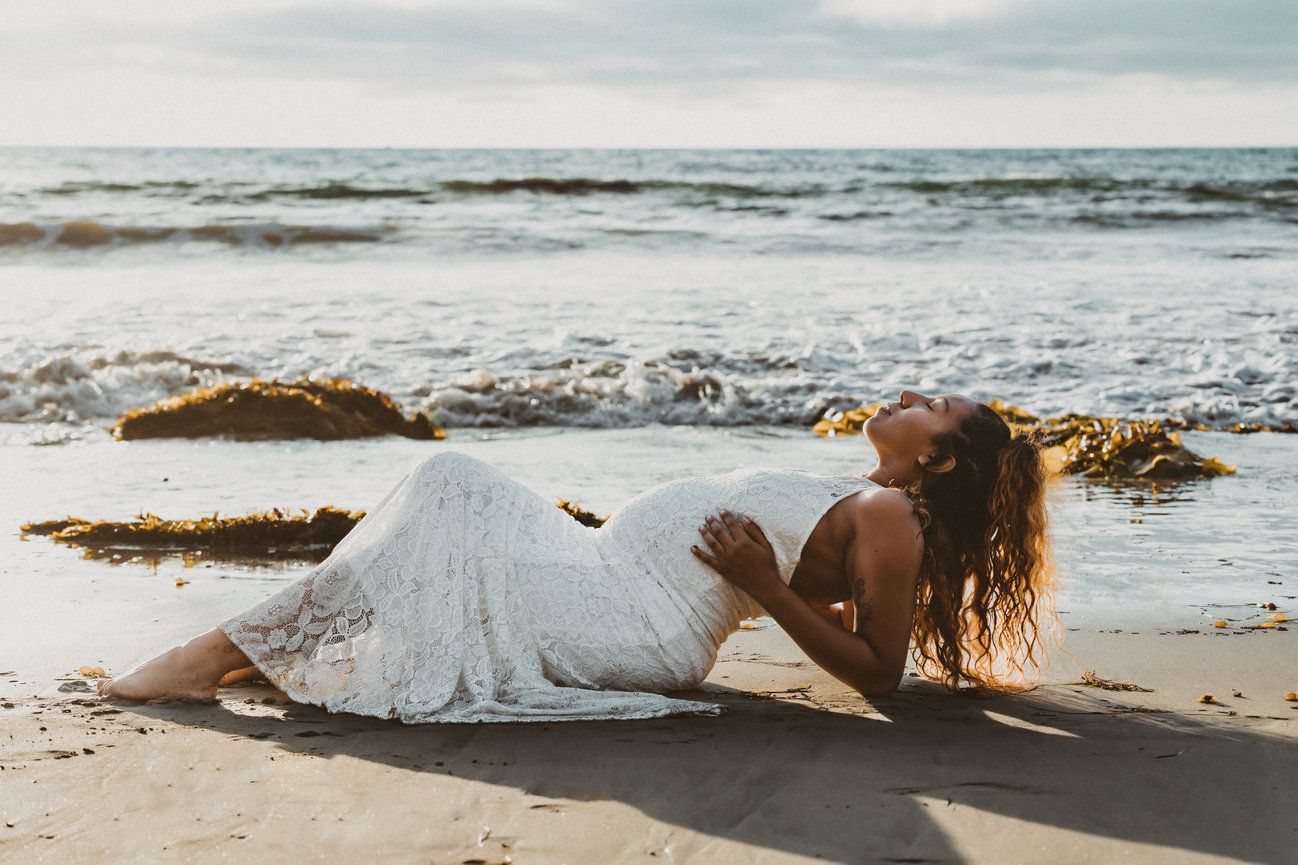 Person in white dress lying on beach at sunset with waves crashing behind.
