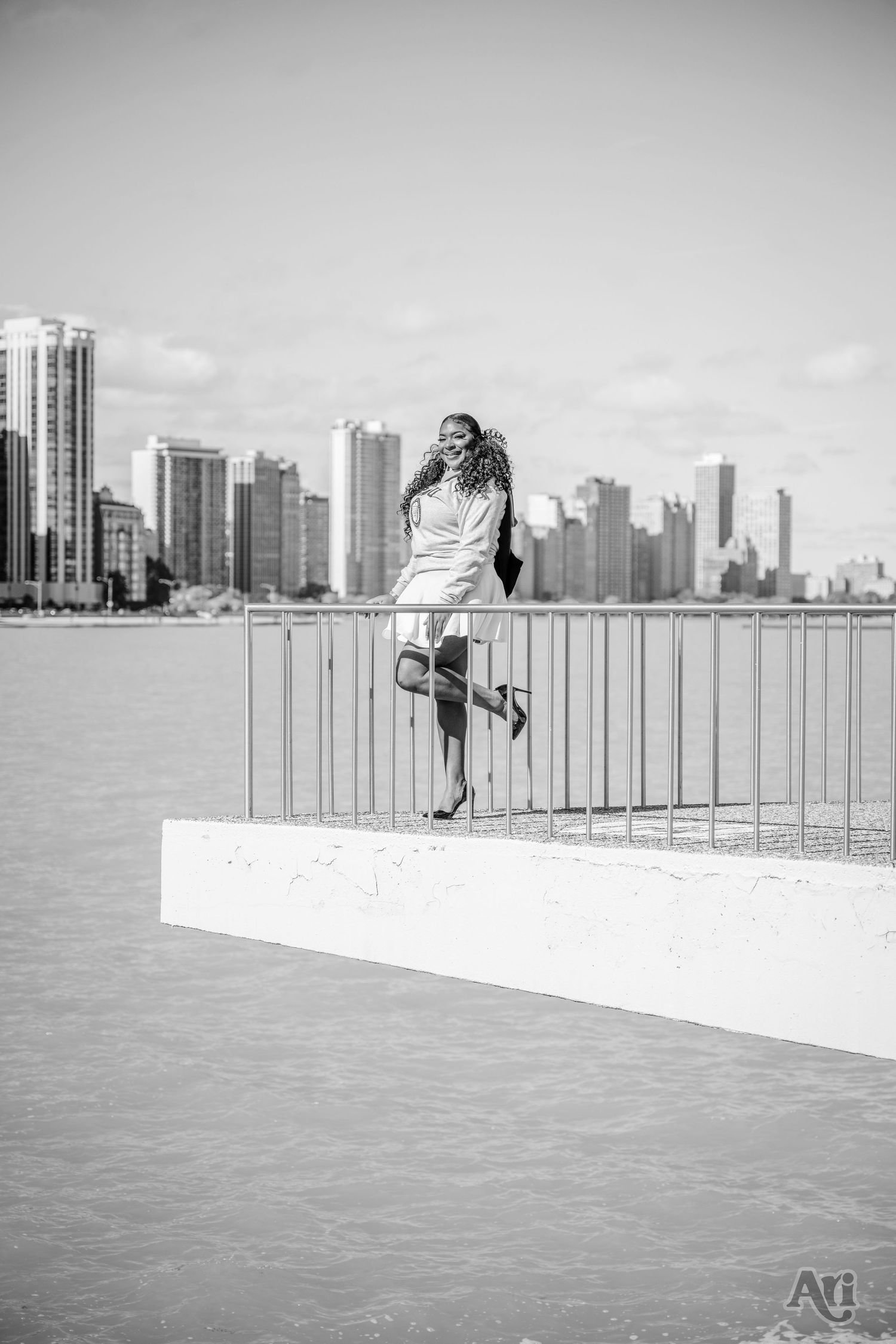 Woman in white dress poses by waterfront with city skyline in background.