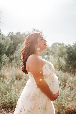 A pregnant woman in a floral white dress stands in a sunlit meadow with lens flares creating a dreamy atmosphere.