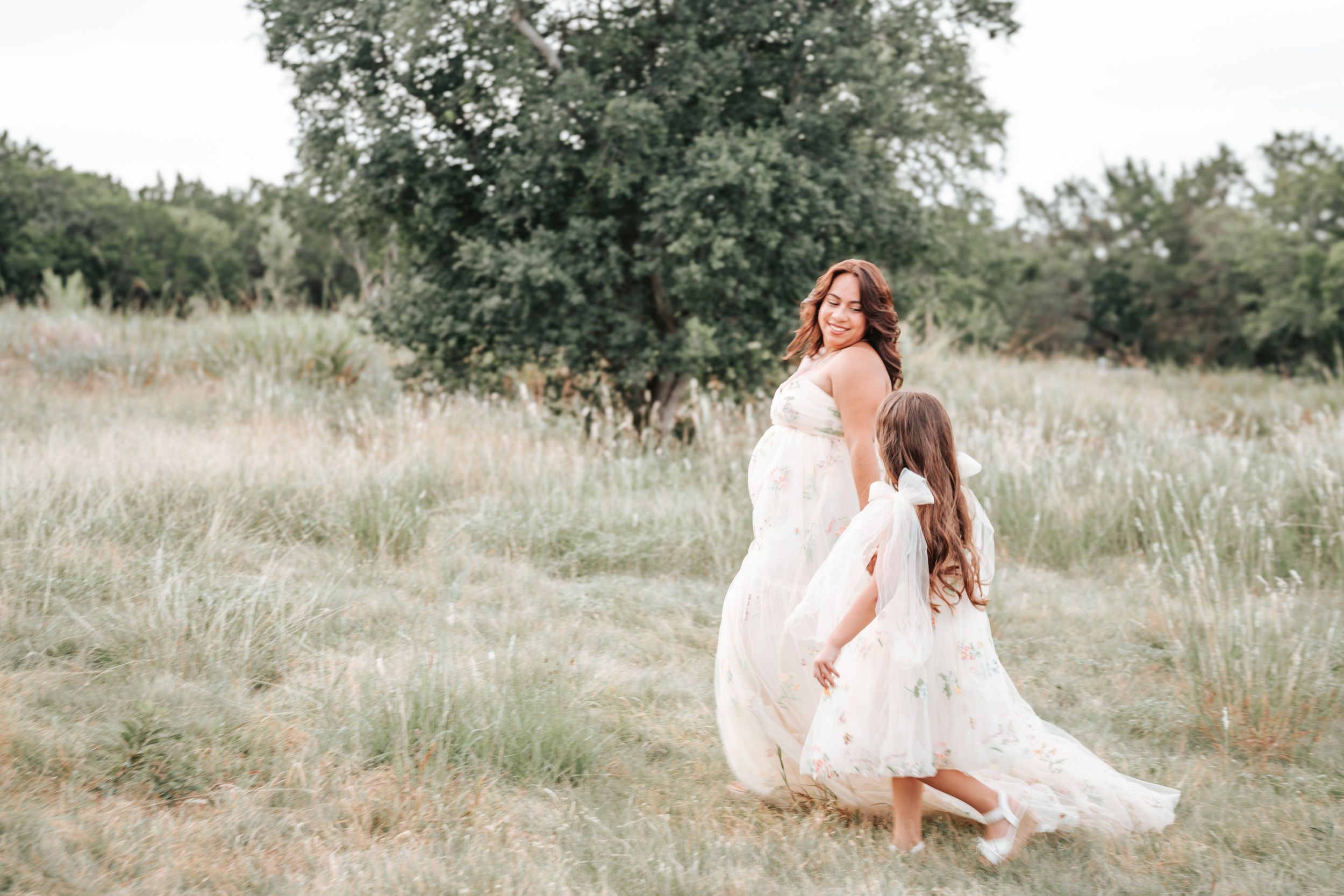 Two people in flowing white dresses dance together in a sunlit meadow with tall grass and trees in the background.