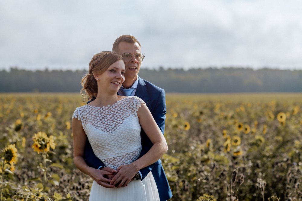 A couple embraces in a sunflower field on their wedding day, with the bride wearing a delicate lace dress.