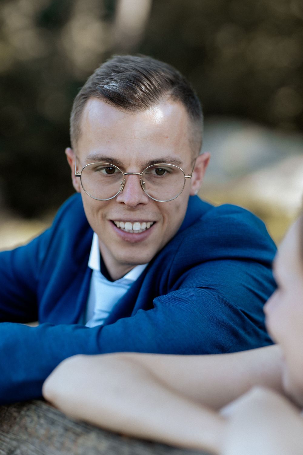 A person wearing glasses and a blue hoodie smiles while relaxing outdoors.