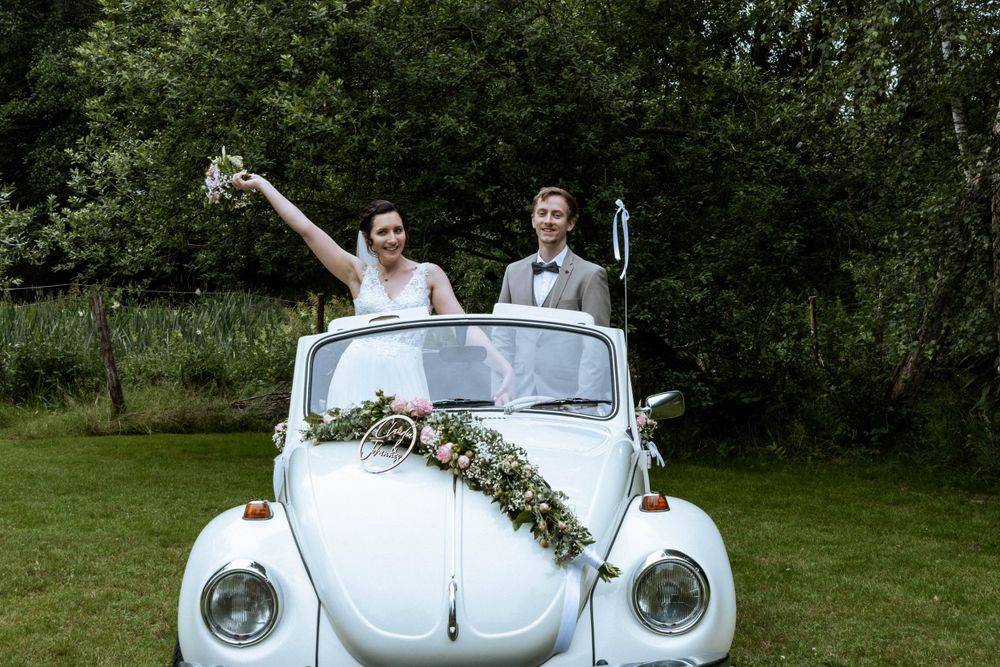 A vintage white Volkswagen Beetle decorated with floral garlands serves as a wedding car on a green lawn.