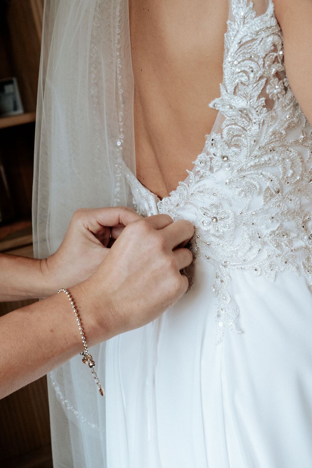Hands fasten delicate lace and beading on a white wedding dress bodice with flowing veil detail.