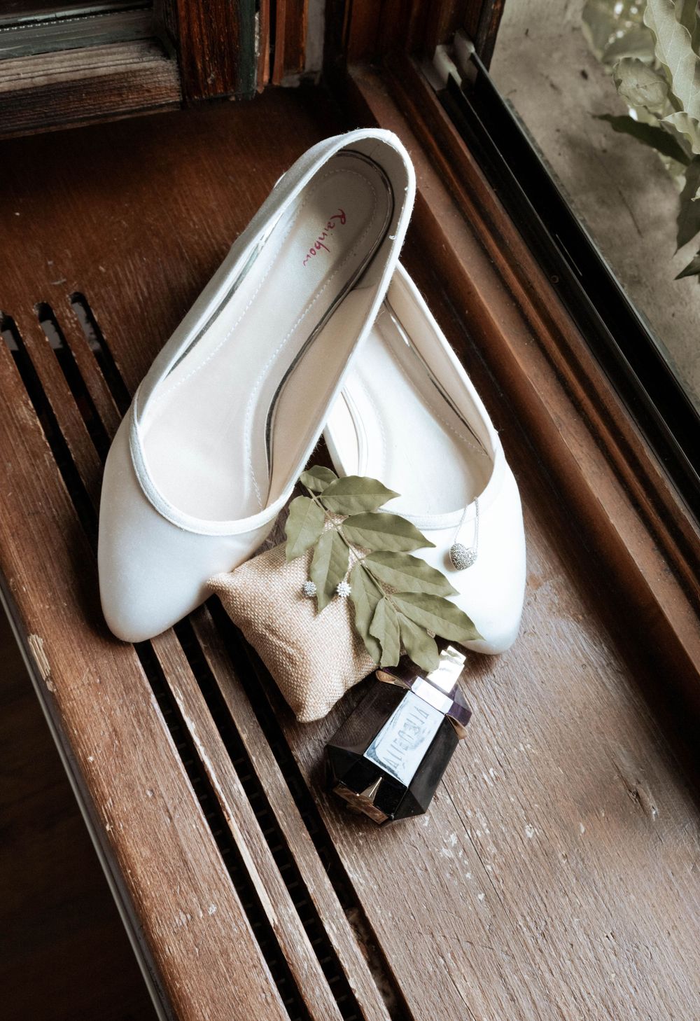 White ballet flats with a small perfume bottle and dried leaves arranged on a wooden surface.