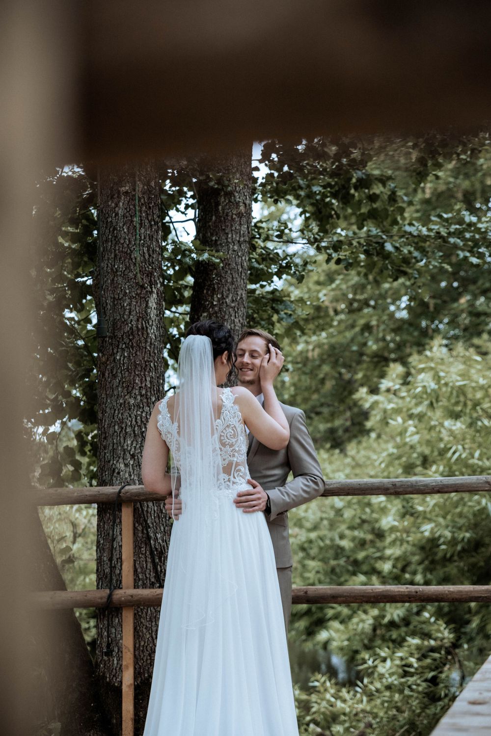 A couple embraces on a wooden deck in the woods, with the bride wearing a white lace gown and flowing veil.