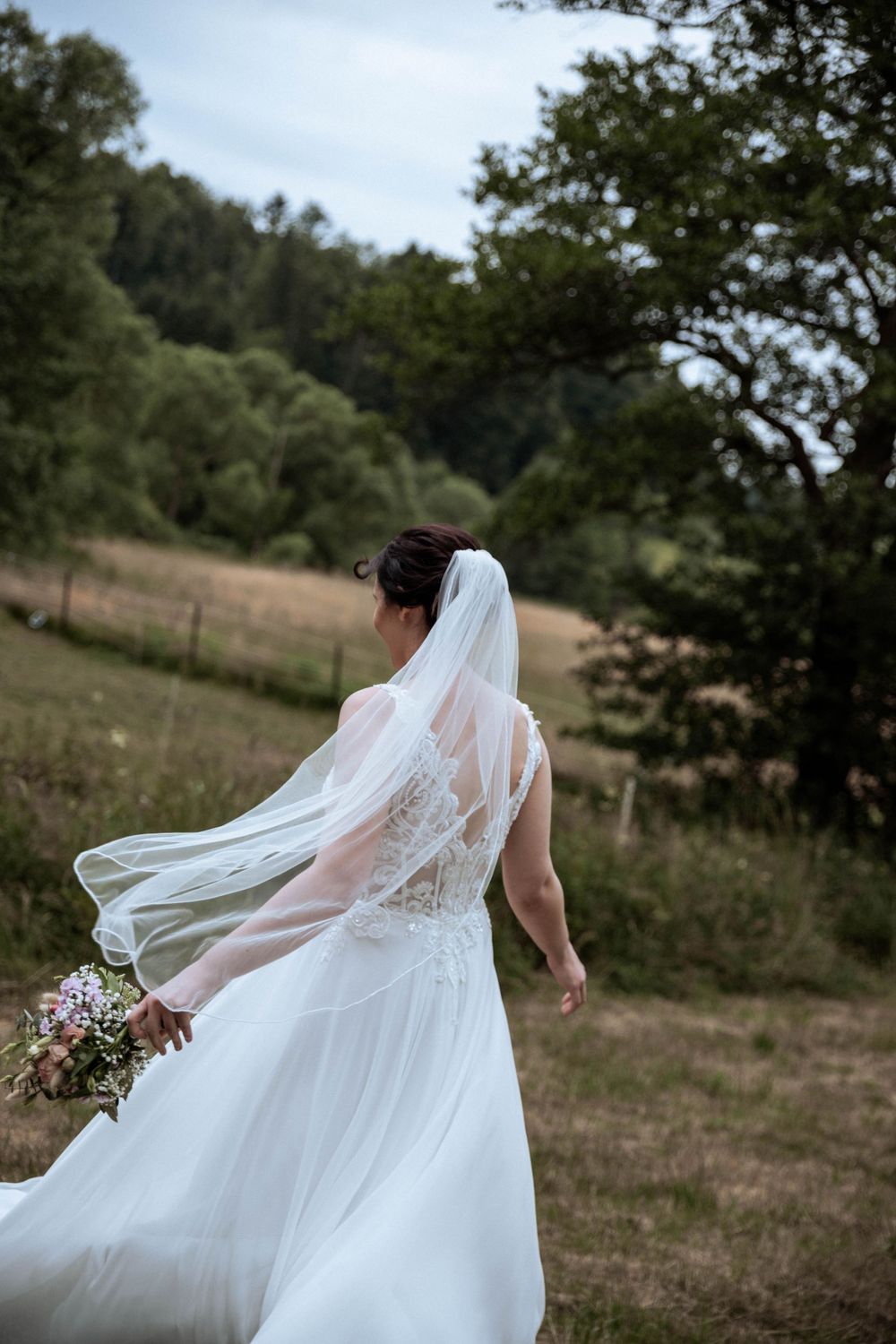 A bride in a flowing white lace dress and veil holding a bouquet walks through a pastoral field with trees in background.