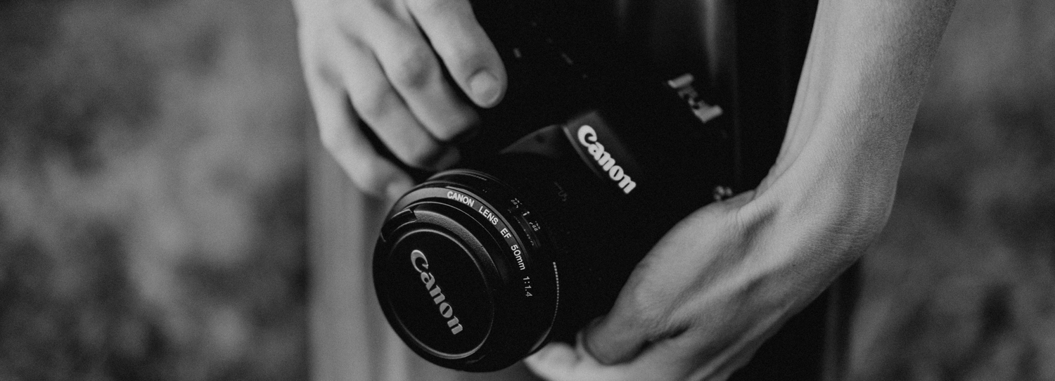 A person holds a black Canon DSLR camera with lens against a blurred dark background.