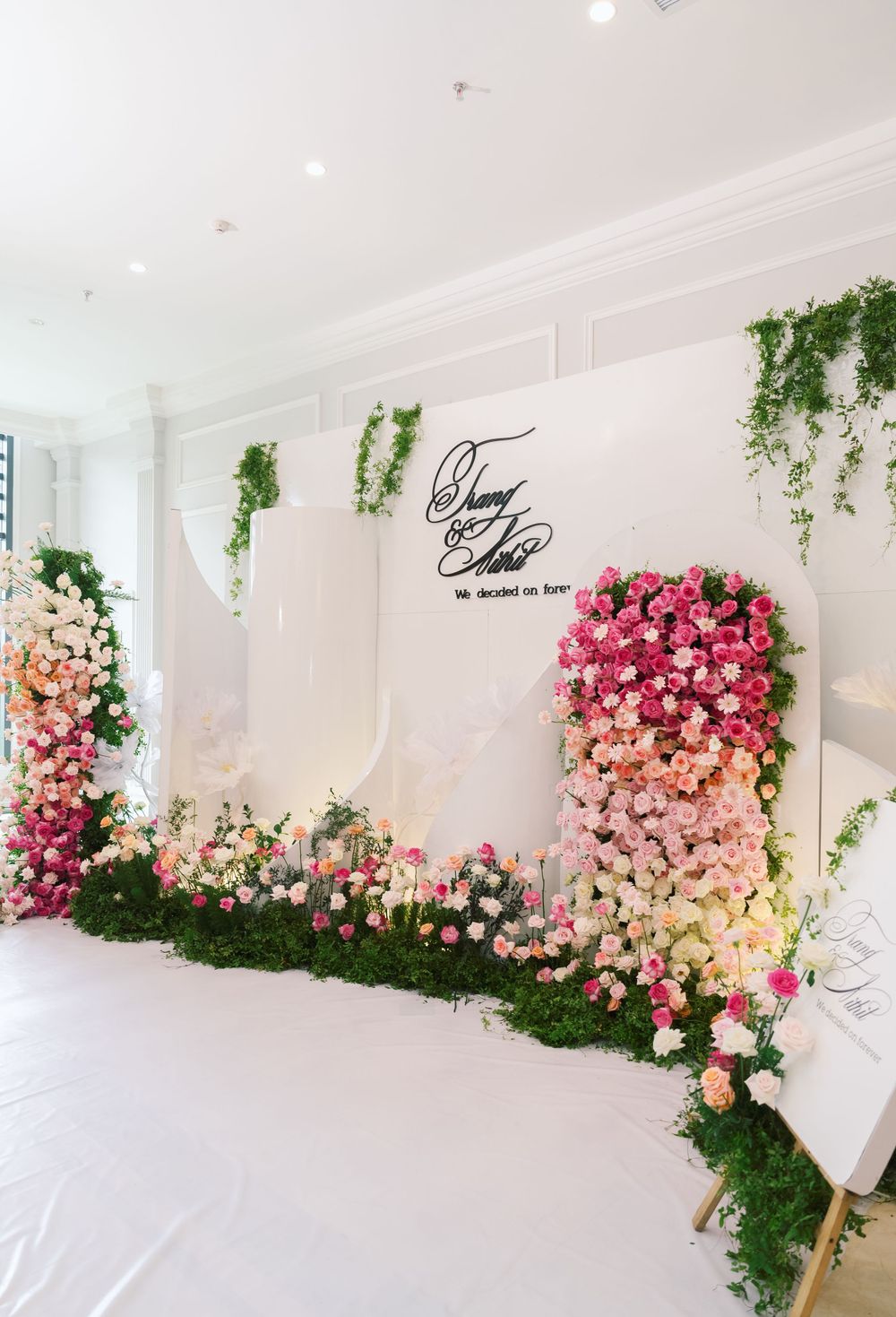 Wedding photo backdrop featuring ombre pink roses and cascading greenery against white wall.