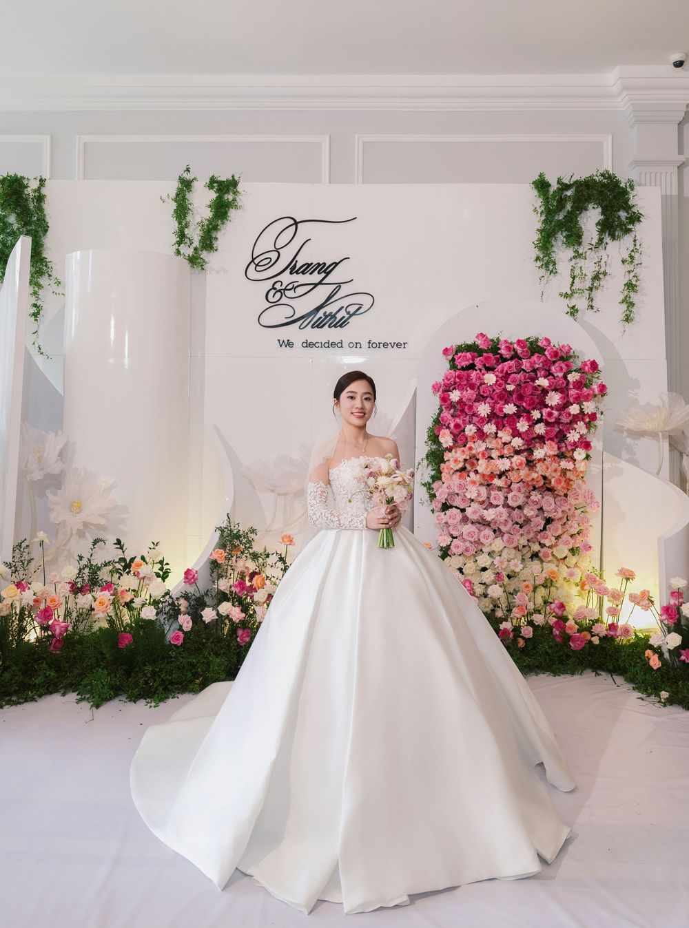Bride in classic white ballgown poses against floral backdrop with pink roses and greenery.
