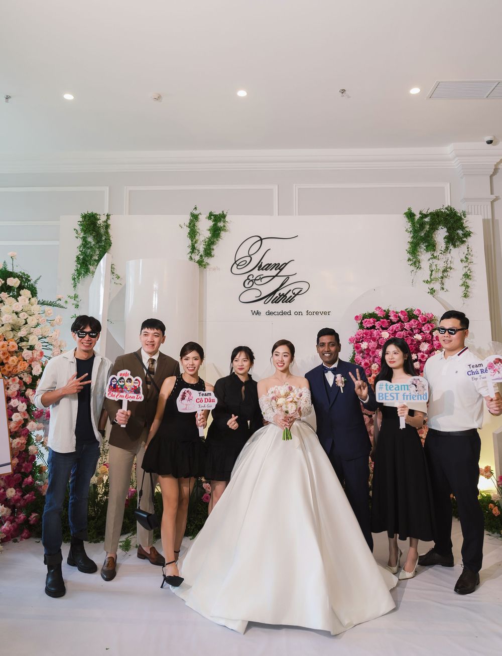 Wedding party group photo in front of floral wall backdrop during elegant indoor celebration.