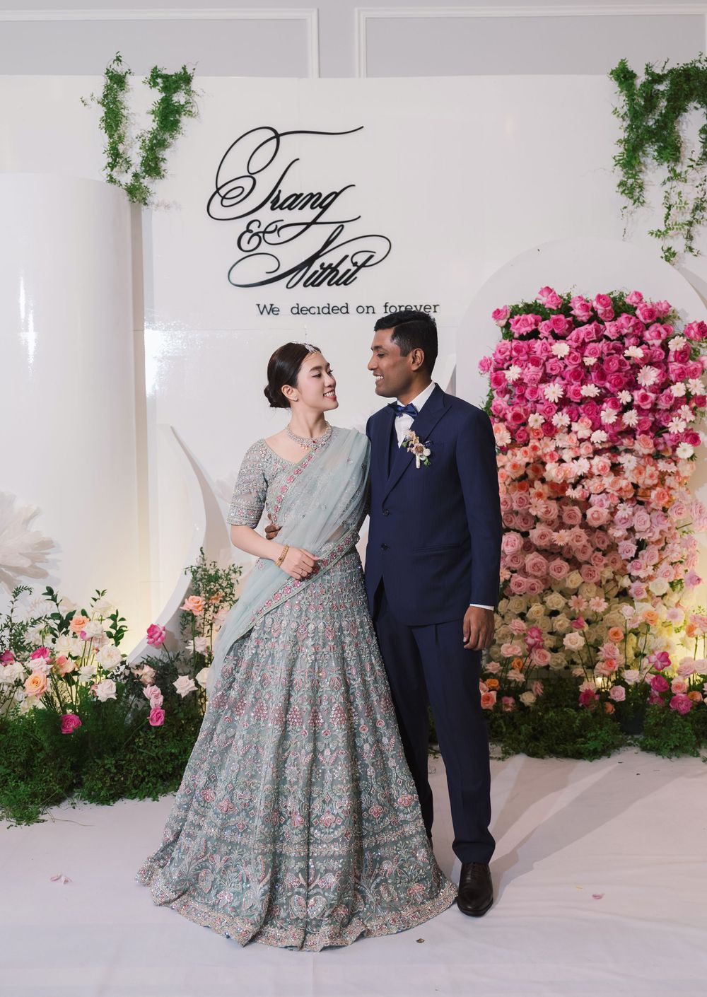 Wedding couple poses against floral wall backdrop with pink ombre roses and greenery.