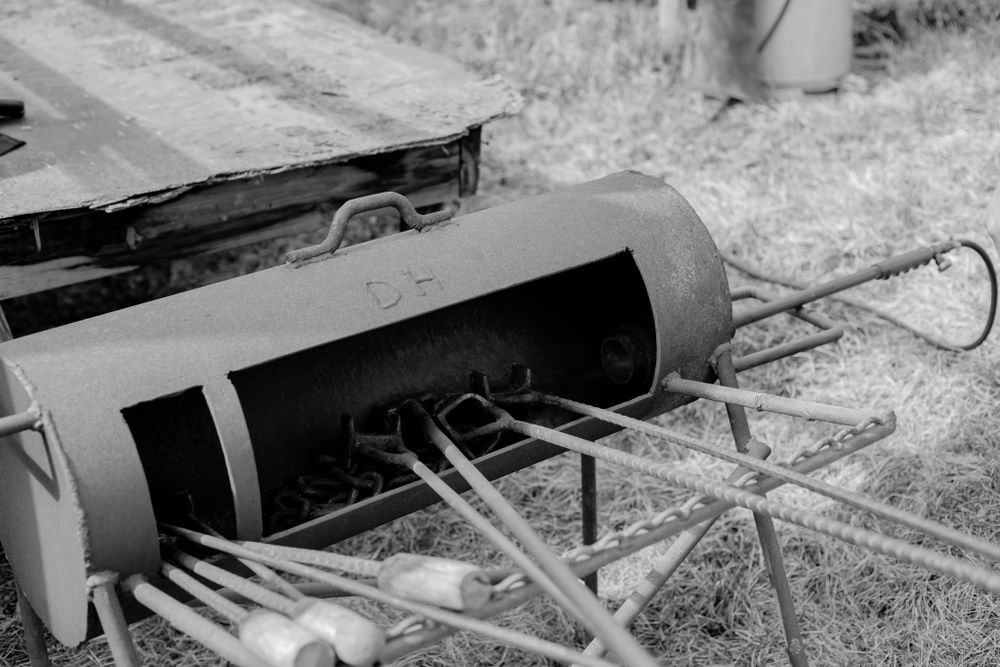 Vintage farm equipment with cylindrical roller and metal tines rests on grass in black and white.