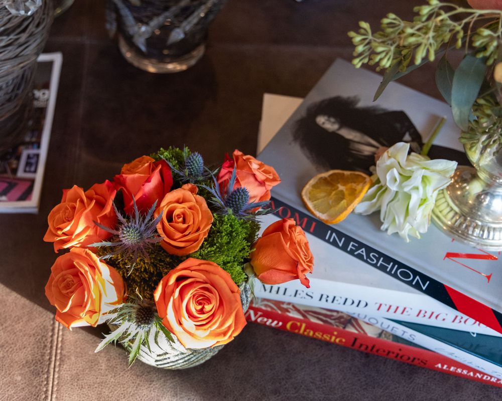 Vibrant orange roses arranged with greenery on a dark surface next to books and reading materials.