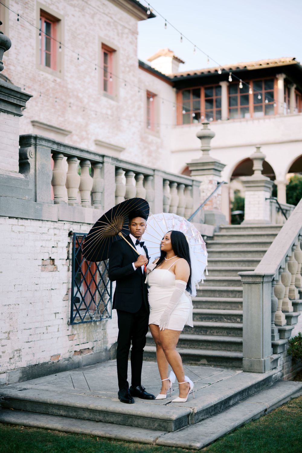 Stylish couple with umbrellas pose on elegant stone staircase of historic building.