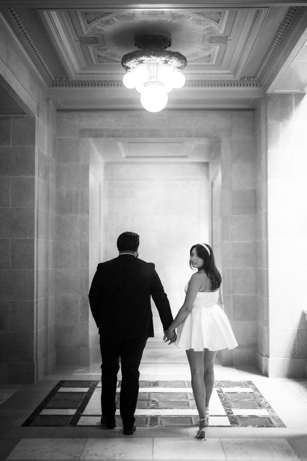 Black and white photo of couple walking hand in hand down an elegant hallway with chandelier.