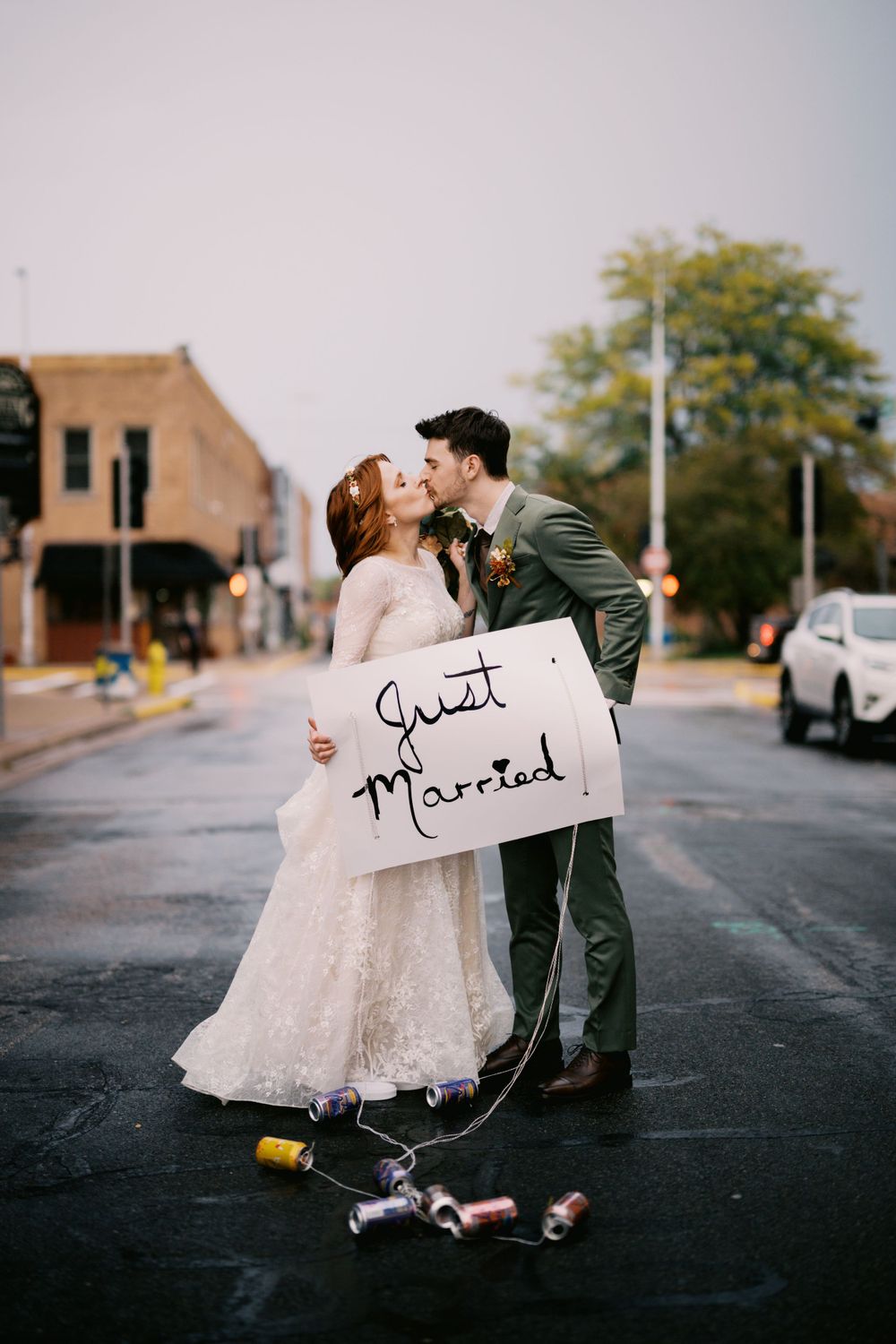 Newlyweds share a romantic kiss on a city street while holding a 'Just Married' sign with cans trailing behind.