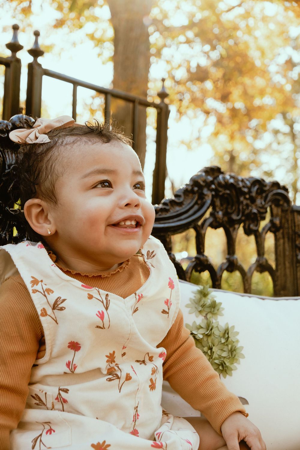 Smiling toddler in floral dress and orange sweater sitting on ornate vintage chair outdoors.