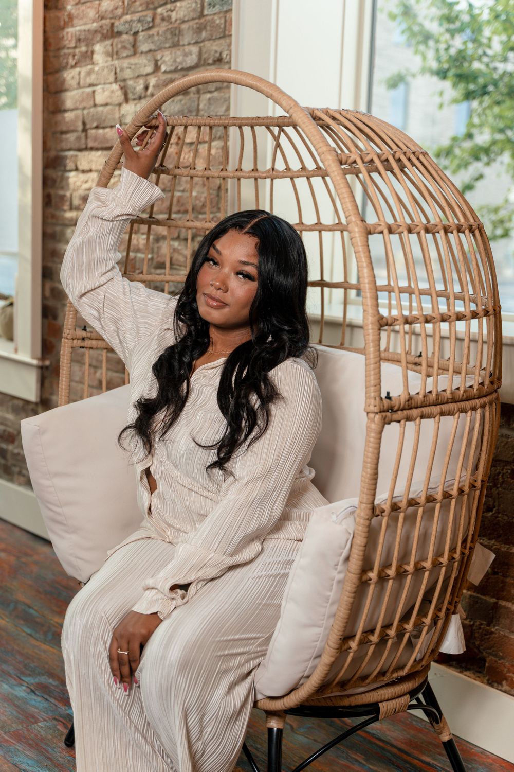 Person relaxing in cozy rattan hanging chair wearing cream knit loungewear against exposed brick wall.