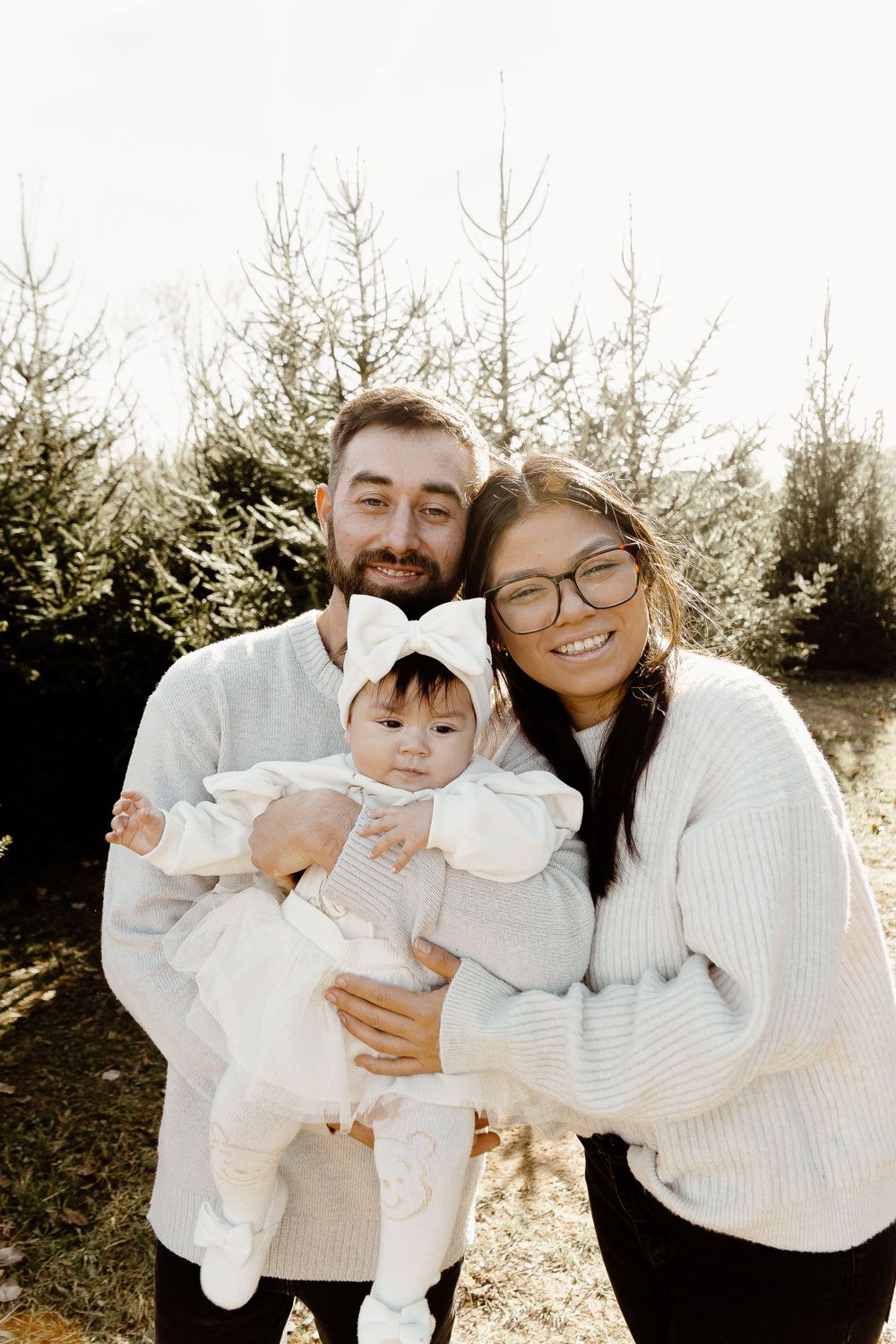 Winter family portrait with evergreen trees in background wearing cozy white sweaters.