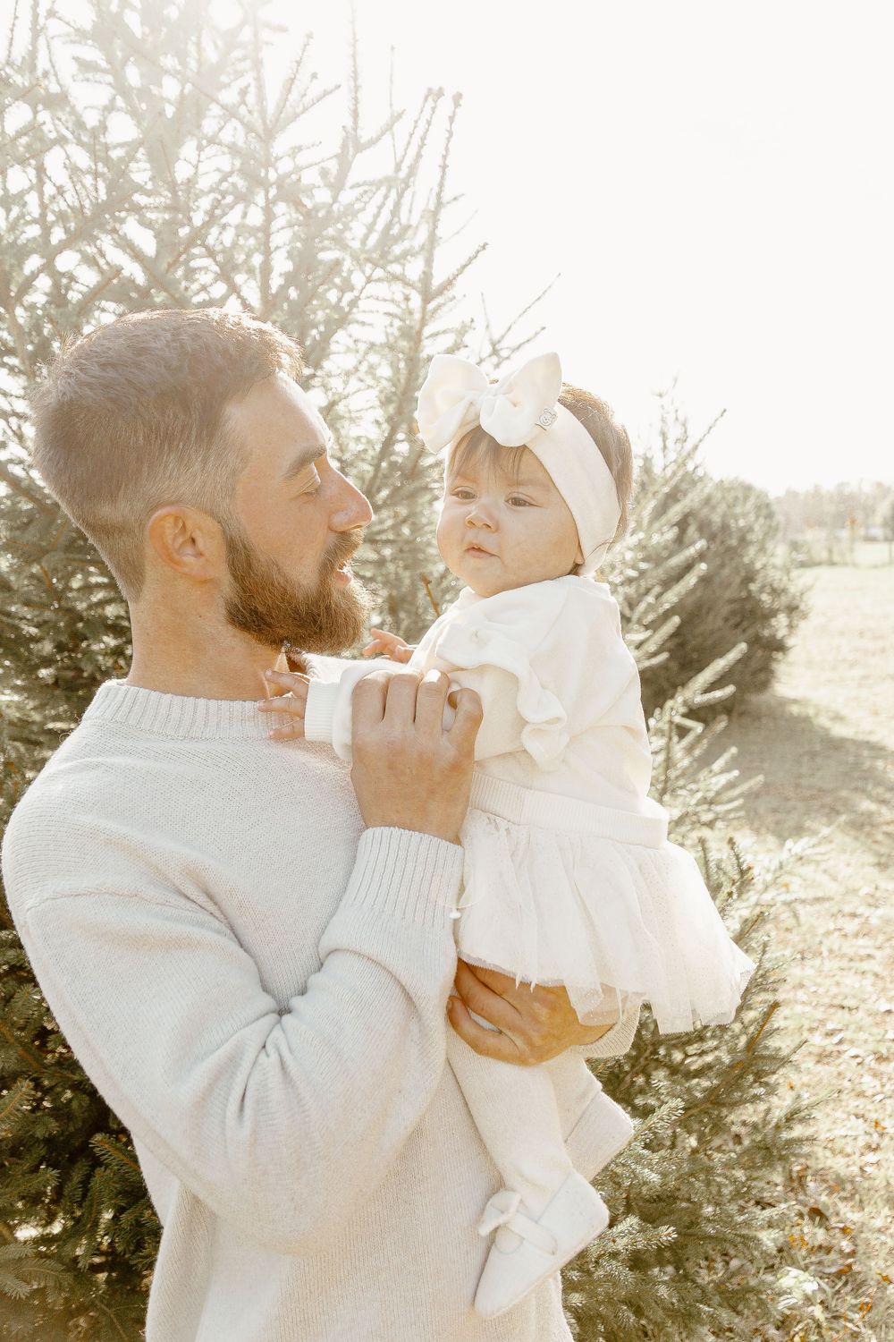 Tender moment between parent and baby in white outfits against evergreen backdrop.