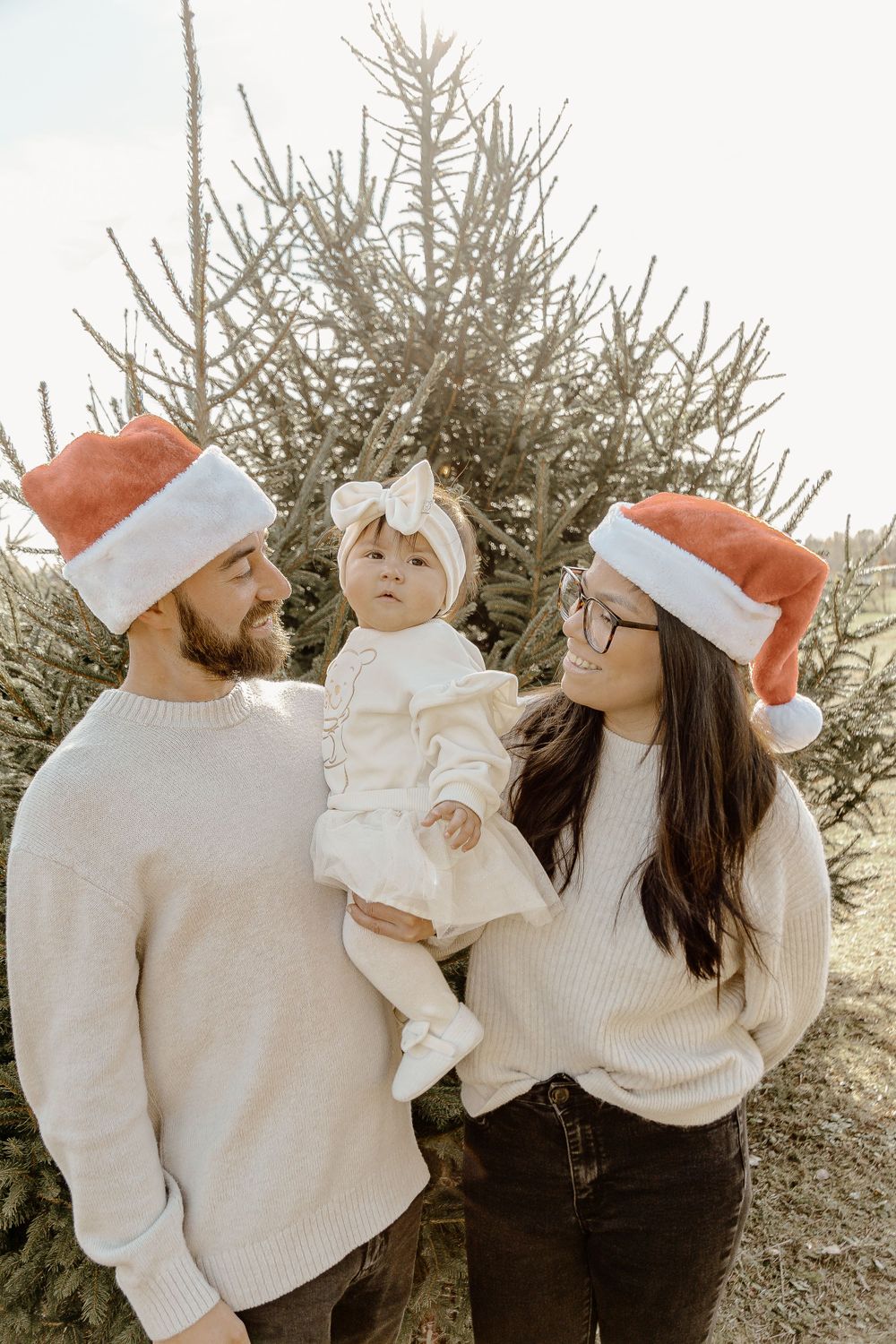 Holiday family photo with Santa hats and white sweaters by Christmas tree.