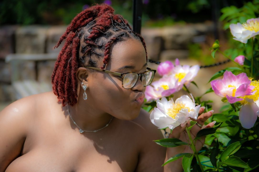 Woman with red locs and glasses smelling pink and white roses in a garden.