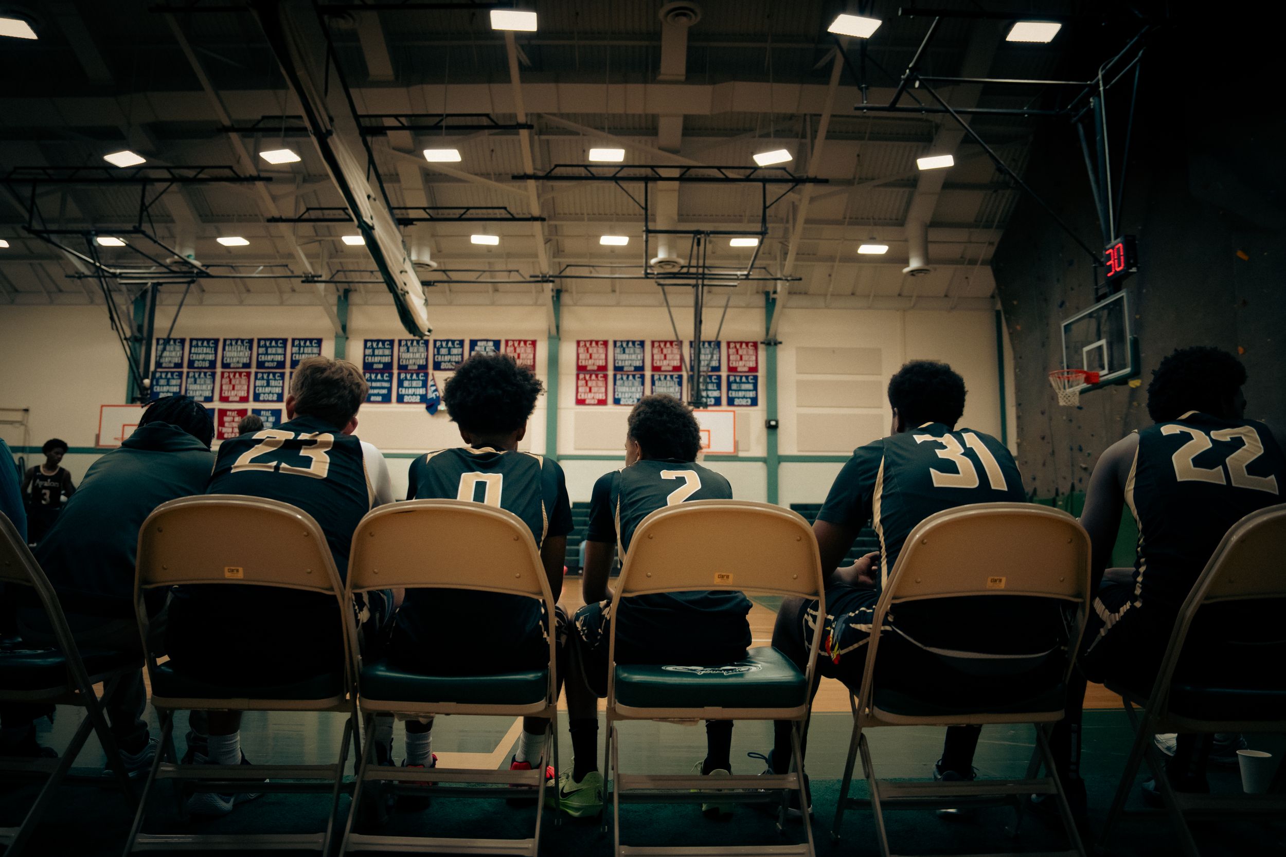 Basketball team sitting on bench chairs viewed from behind in indoor gymnasium.