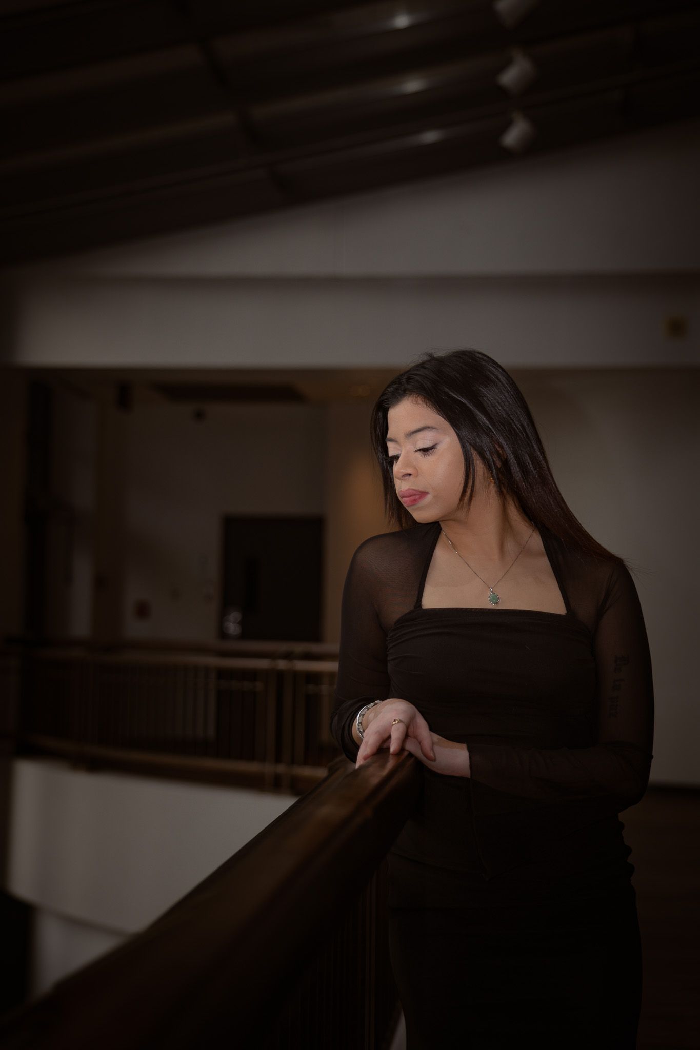 Young woman in a black outfit leans on a wooden railing in a dimly lit interior space.