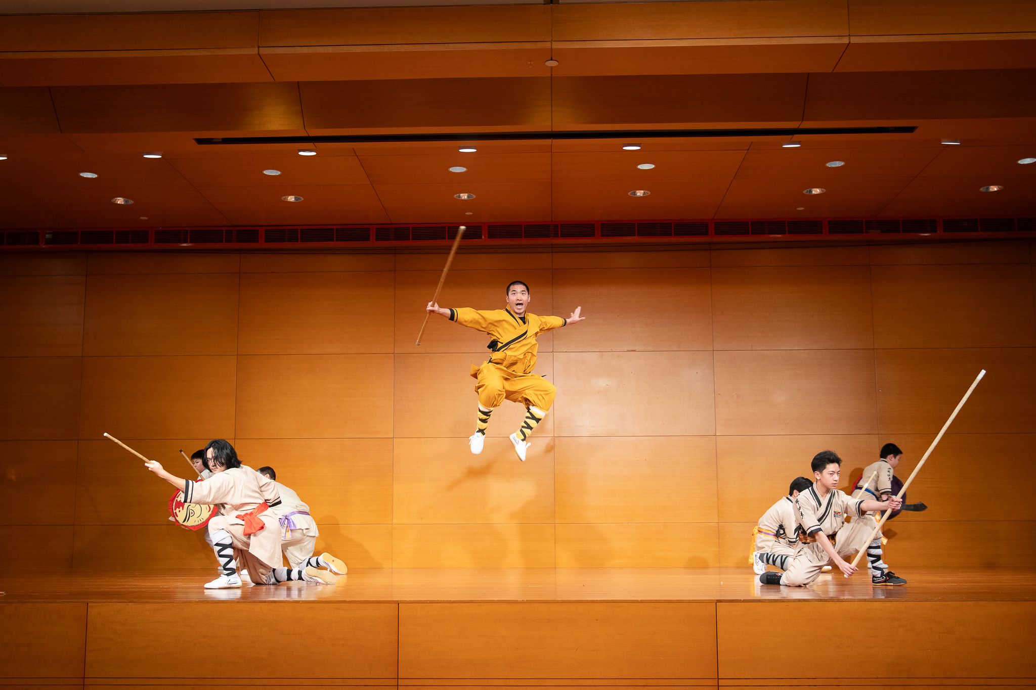 Martial arts performer leaps high above other performers holding staffs on a wooden stage.