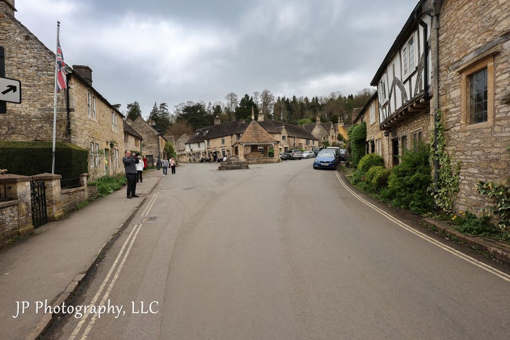 Quiet village street lined with traditional Cotswold stone buildings under cloudy skies.