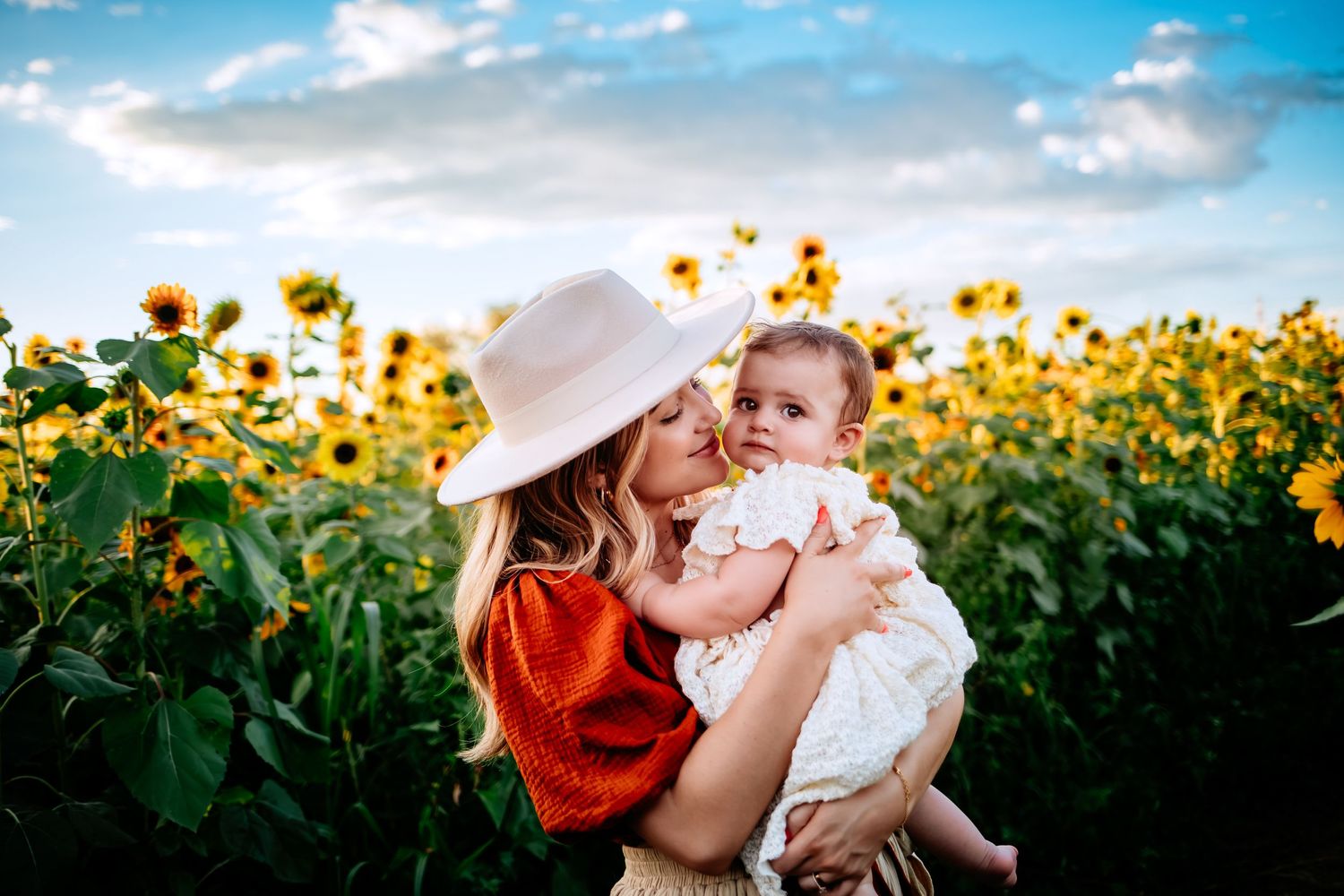 Mother in sun hat embraces baby in white dress against backdrop of blooming sunflower field.