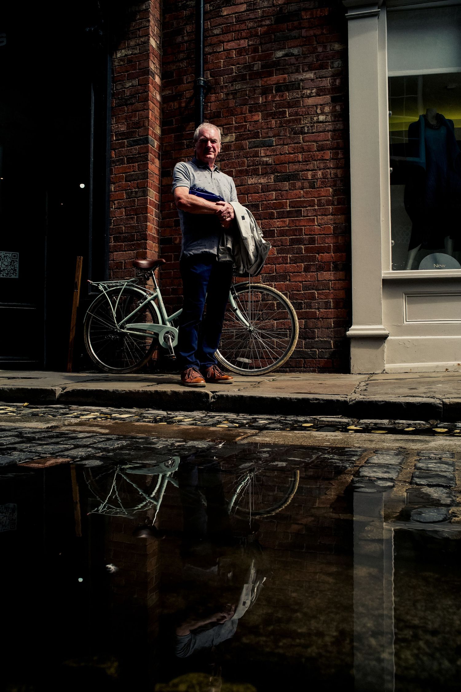 A bicycle leans against a brick wall with a puddle reflecting the scene in an urban street setting.