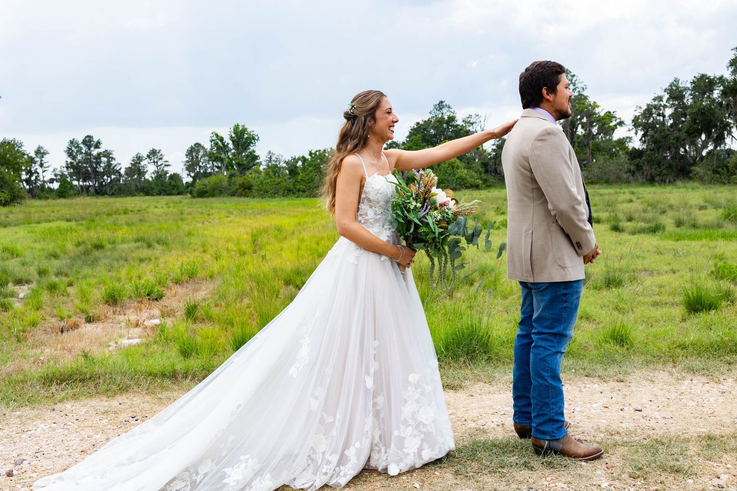 A bride in a white gown touches the shoulder of a groom in a tan jacket outdoors.