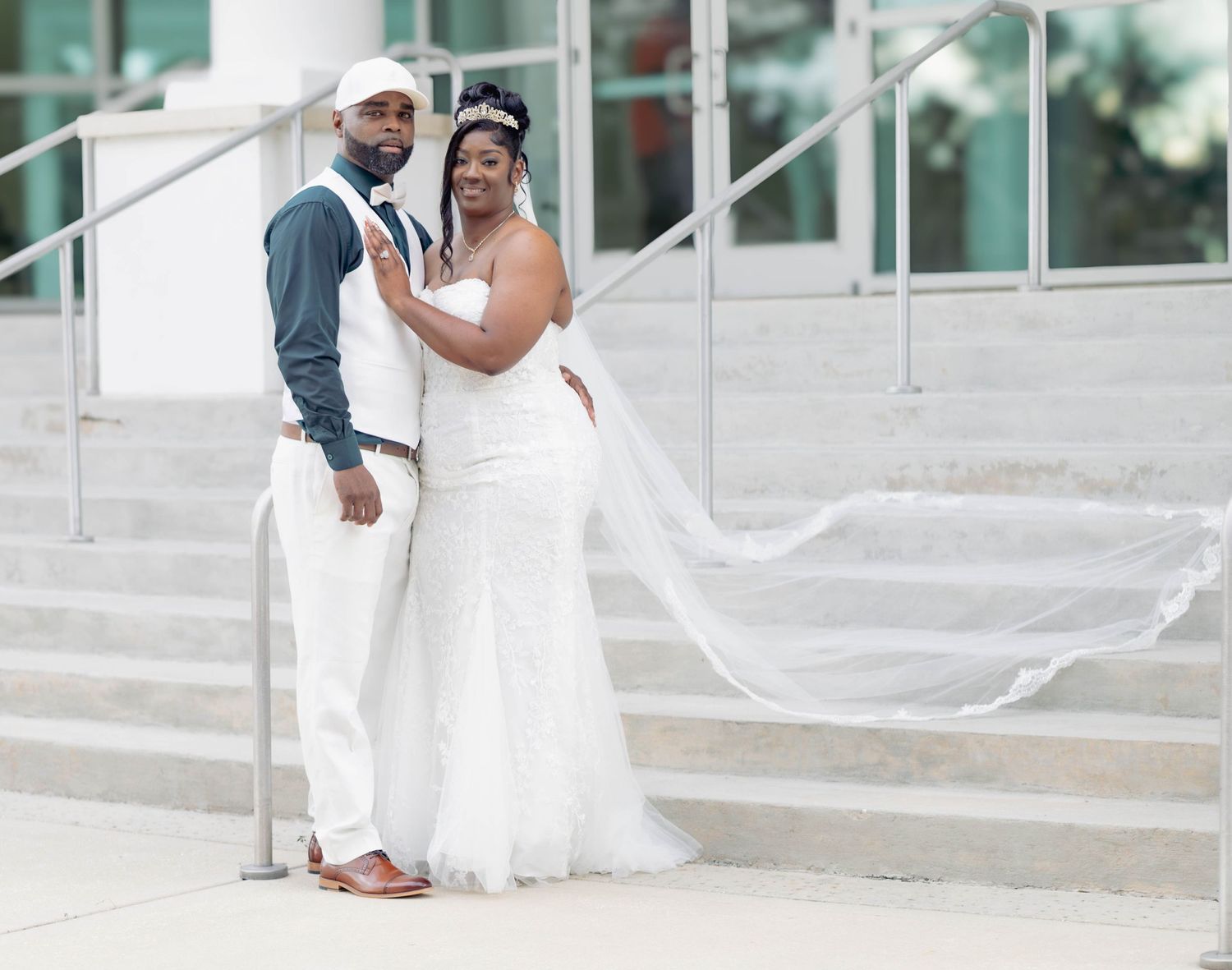 Elegant bride and groom in white wedding attire posing on steps outside a modern building.