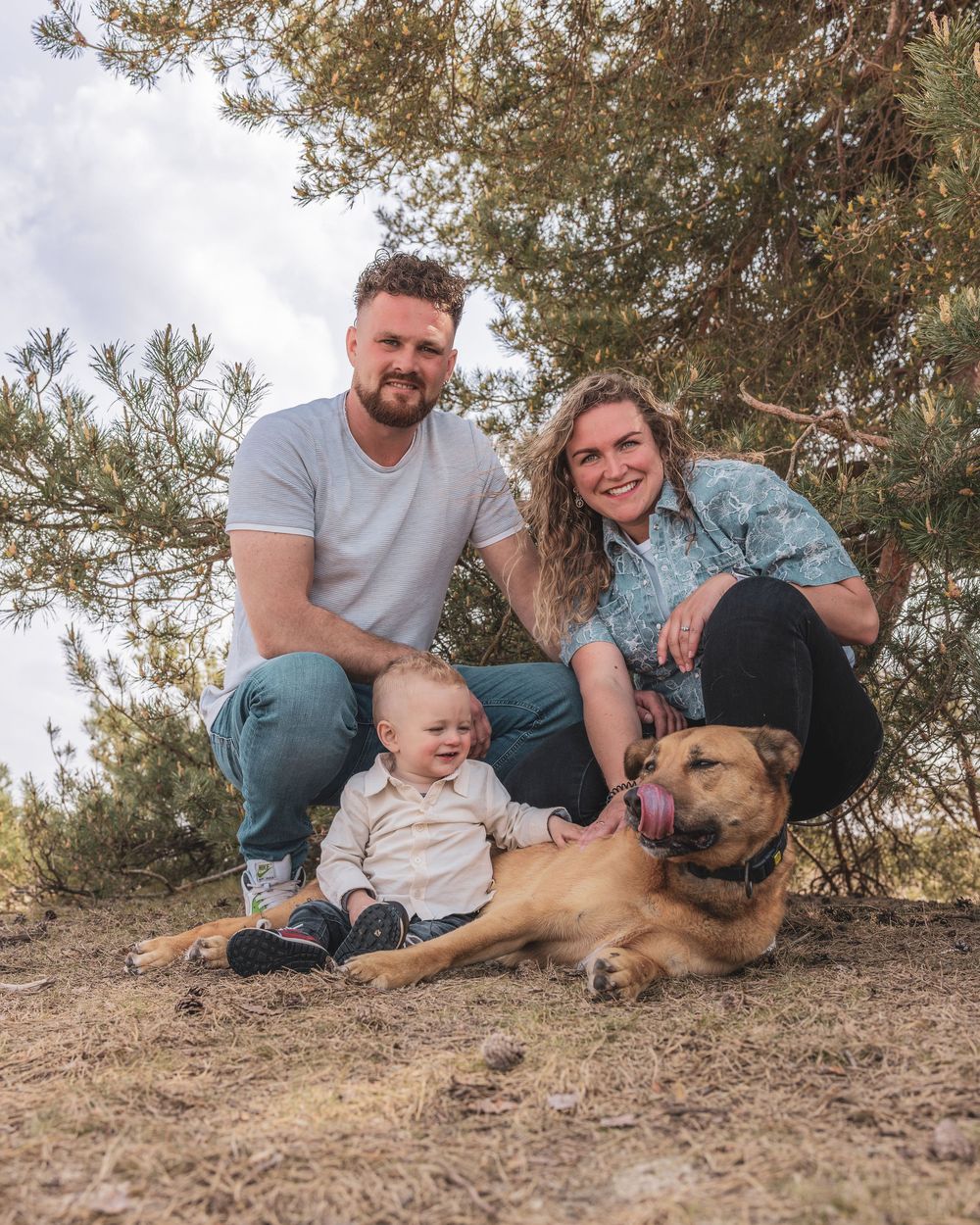 A couple with a baby and a brown dog pose outdoors under a pine tree on a sunny day.