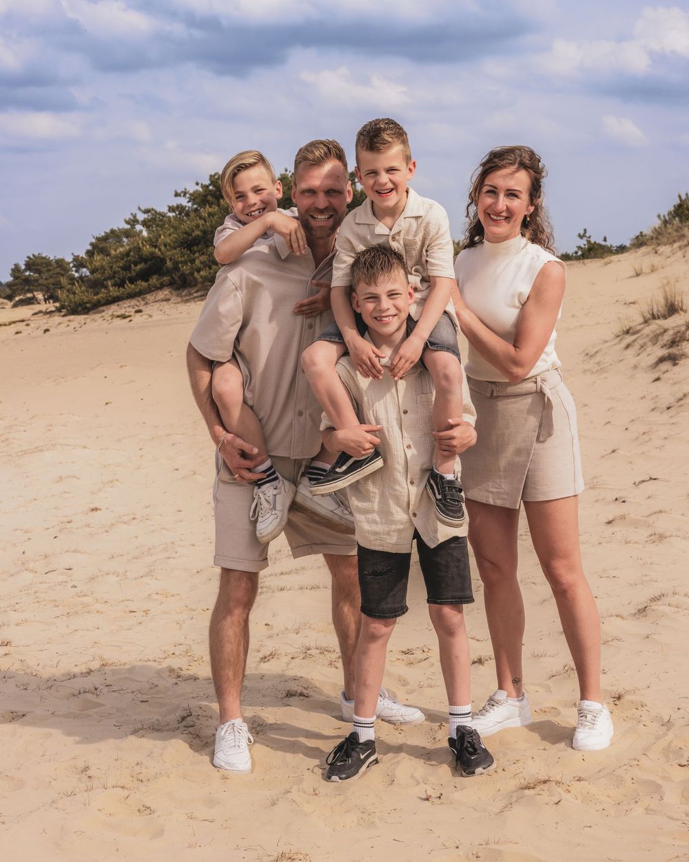 Happy family of five posing together on a sandy beach dune on a cloudy day, wearing matching neutral outfits.