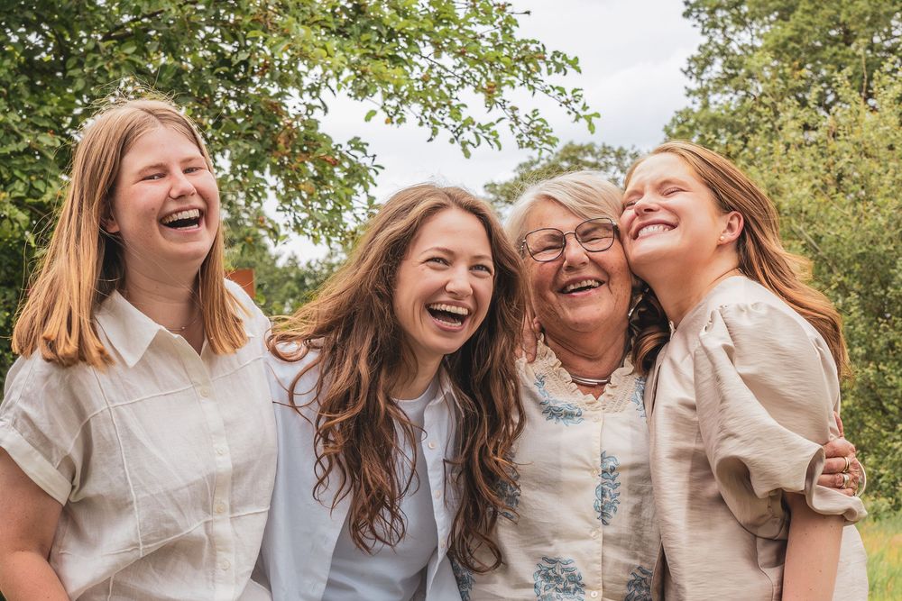 Four women laughing joyfully together outdoors, surrounded by lush green trees in a warm family portrait.